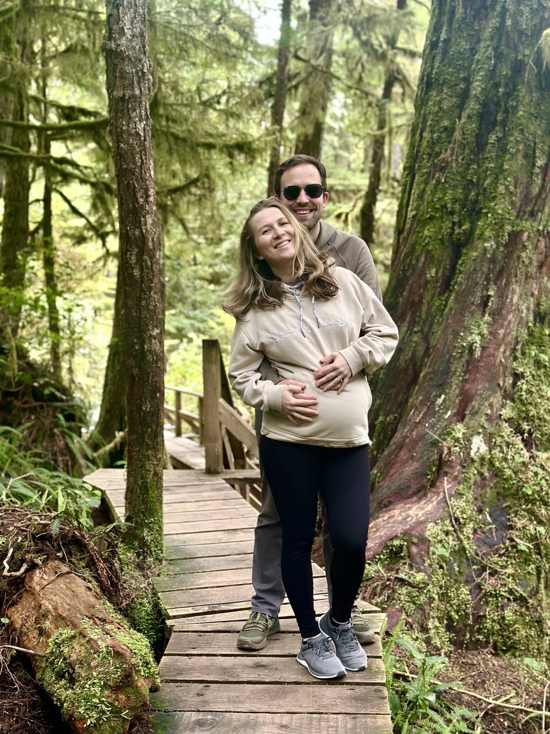 Hosts Anna and Vrat smiling on a wooden boardwalk in the forest while hiking in Cape Breton, Nova Scotia